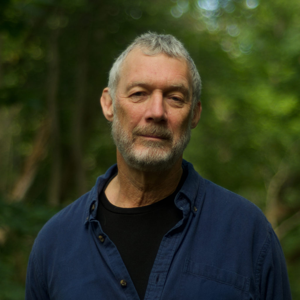a colour photograph of a man looking at the camera. in the background a blur of green trees.