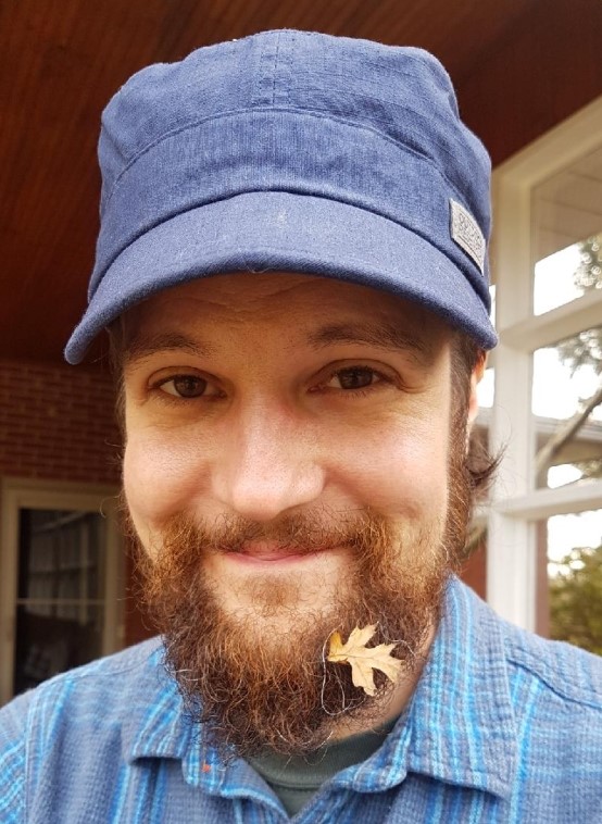 a colour photo of a man smiling with a small leaf stuck to his beard.